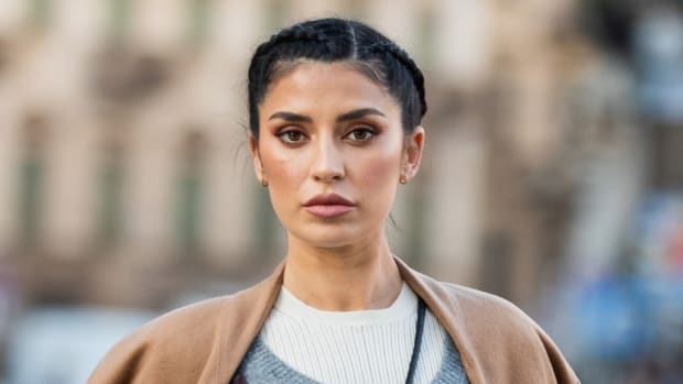 Nicole Williams English poses on the street in a white sweater and a tan coat. She wears her black hair in a braided updo.